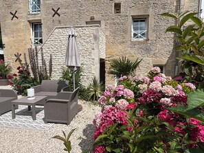 Un bâtiment en pierre doté d’une terrasse extérieure, d’un patio couvert et d’un jardin agrémenté de fleurs roses.