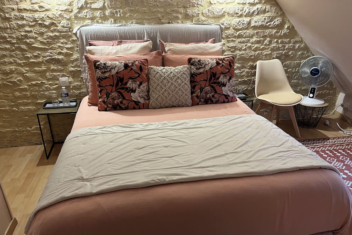 Une chambre avec un mur en pierre, un lit recouvert d’une couverture rose, plusieurs oreillers, une chaise et un ventilateur.