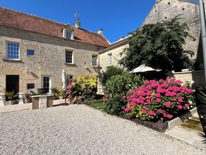 Un bâtiment en pierre avec une cour recouverte de gravier, des plantes en pot et un ciel bleu dégagé.
