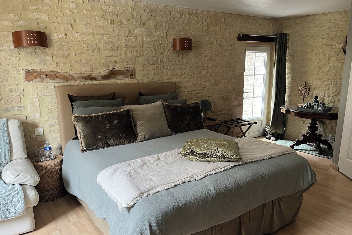 Une chambre avec un mur en pierre, un lit avec de nombreux oreillers, une table de chevet et une fenêtre avec des rideaux.