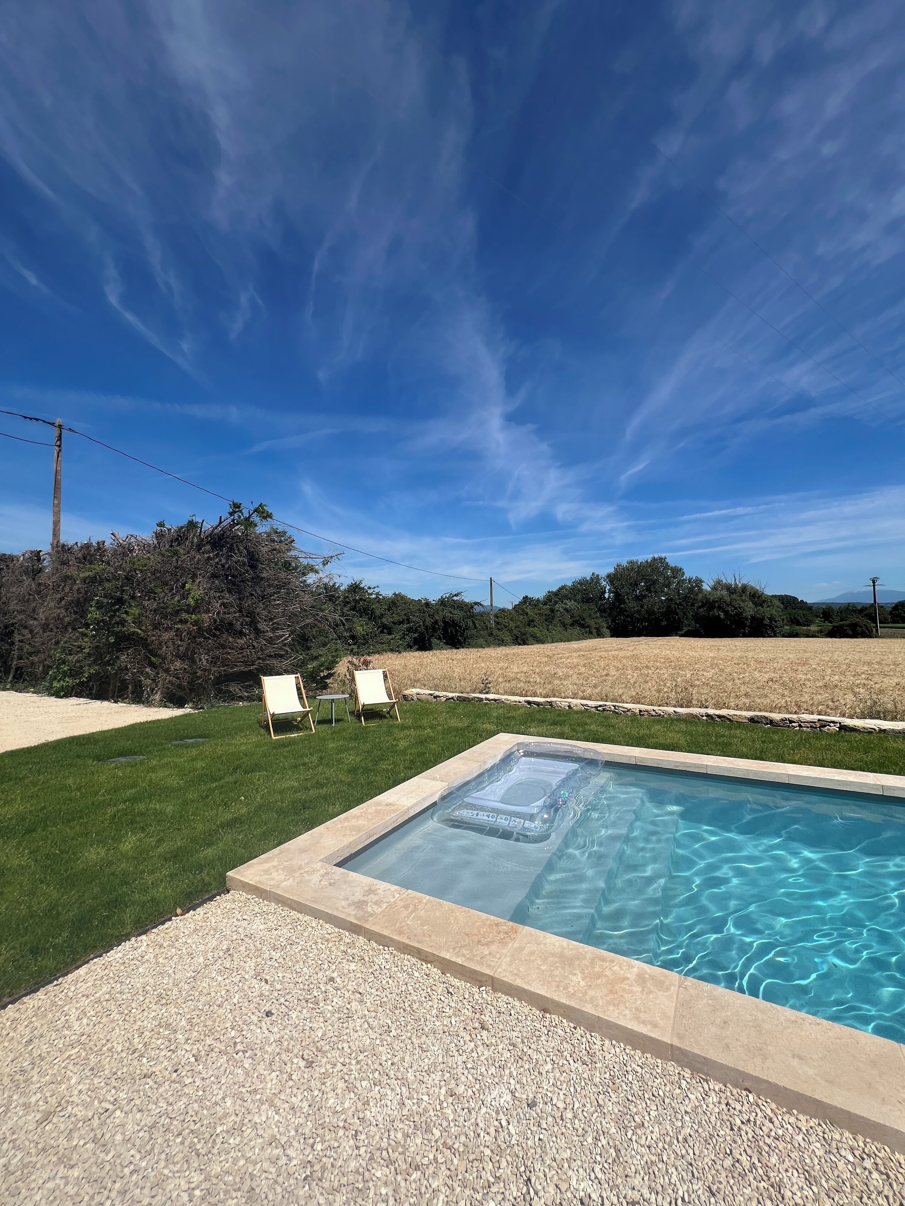 A rectangular hot tub with a control panel, surrounded by a pebble border, set against a backdrop of a grassy lawn and a field.