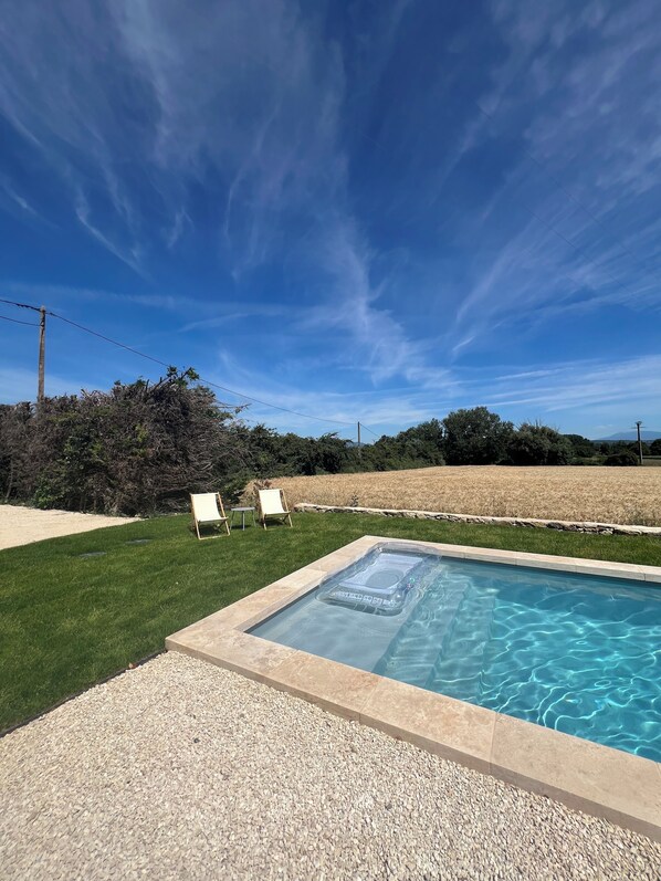A rectangular hot tub with a control panel, surrounded by a pebble border, set against a backdrop of a grassy lawn and a field.