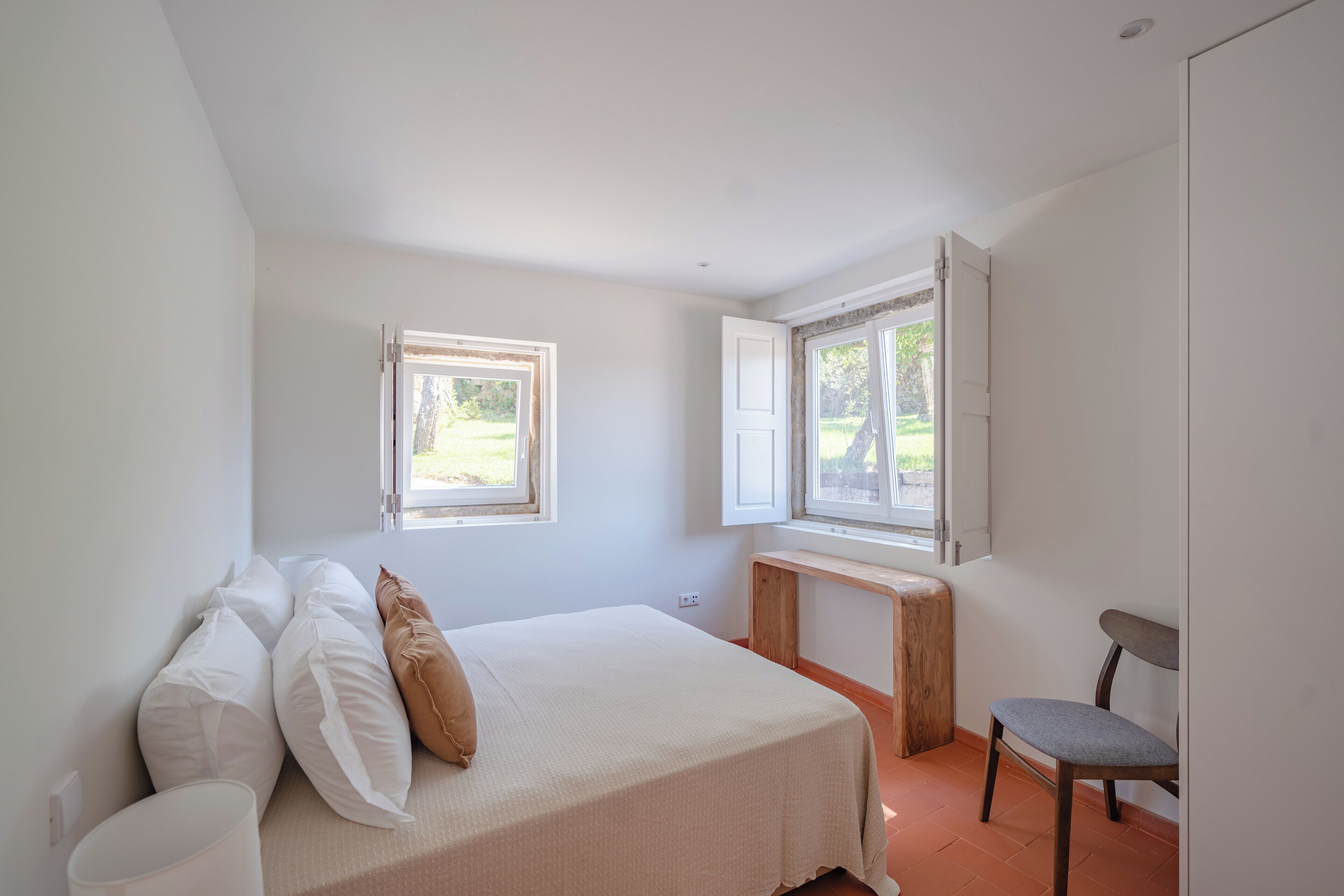 A bedroom with a bed, two windows, a wooden bench, and a chair.