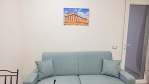 Um sofá azul claro com duas almofadas, uma mesa pequena com toalha de mesa estampada e um quadro de um templo grego antigo na parede.