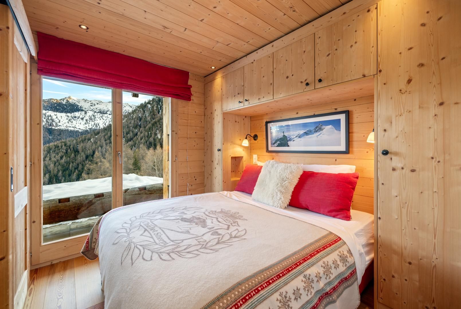 Ein Schlafzimmer in einer Holzhütte mit einem Bett, einem Fenster mit rotem Vorhang, einem gerahmten Bild an der Wand und Blick auf eine verschneite Berglandschaft.