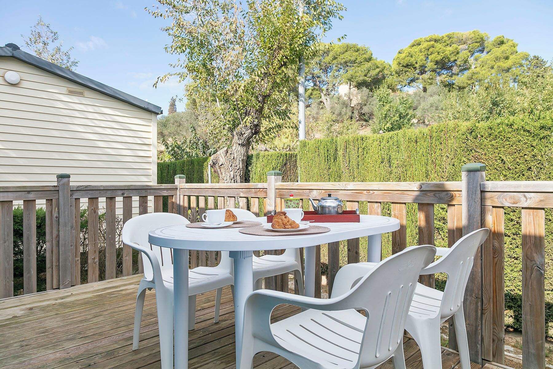 A deck with a round table and white plastic chairs, a teapot, and a croissant on the table.