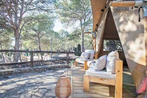 Una terraza de madera al aire libre con un sofá y cojines, rodeada de árboles y un muro de piedra.