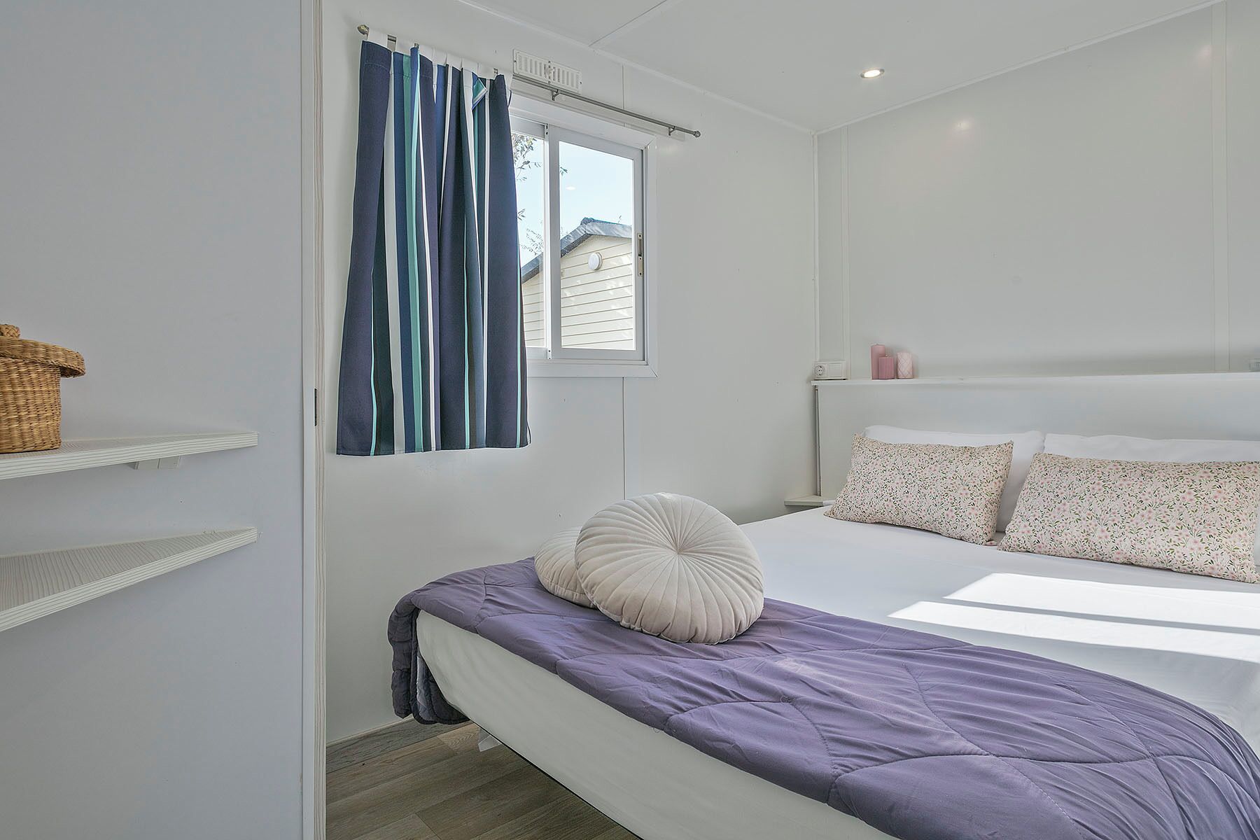 A bedroom with a bed, a window with curtains, a bedside table, and a wall-mounted shelf.