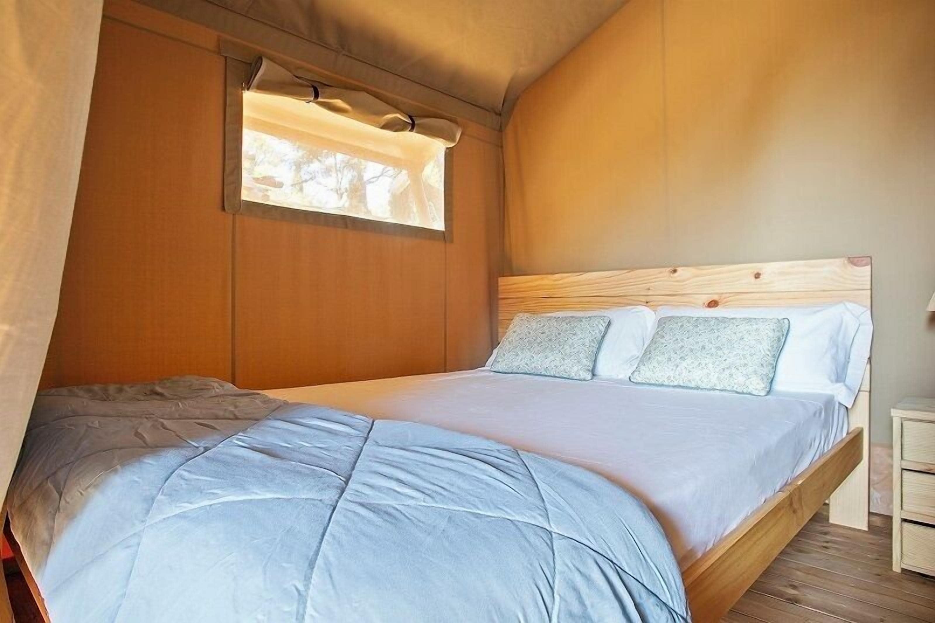 Una cama individual con cabecera de madera, una ventana con cortinas y una mesita de noche.