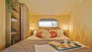 A cozy, well-lit tent-style room with a bed, pillows, a tray with books and pastries, and a shelf with books and a plant. - Camping Castell Montgrí (Torroella de Montgrí)