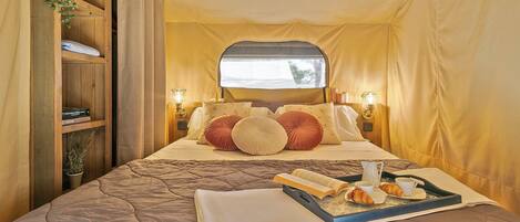A cozy, well-lit tent-style room with a bed, pillows, a tray with books and pastries, and a shelf with books and a plant.