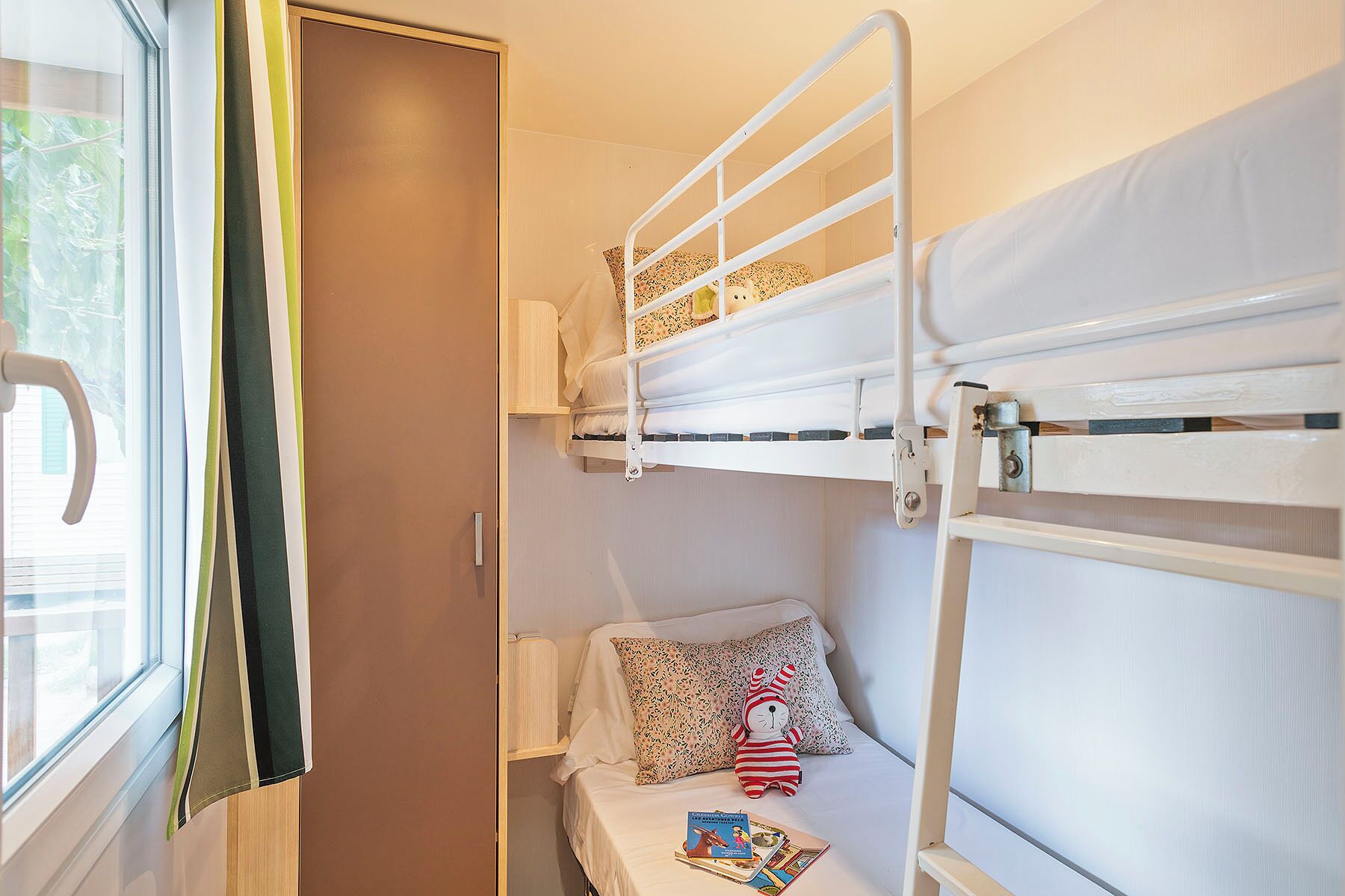 A bunk bed room with a window, a door, and a small bedside table with a pillow and a stuffed toy.