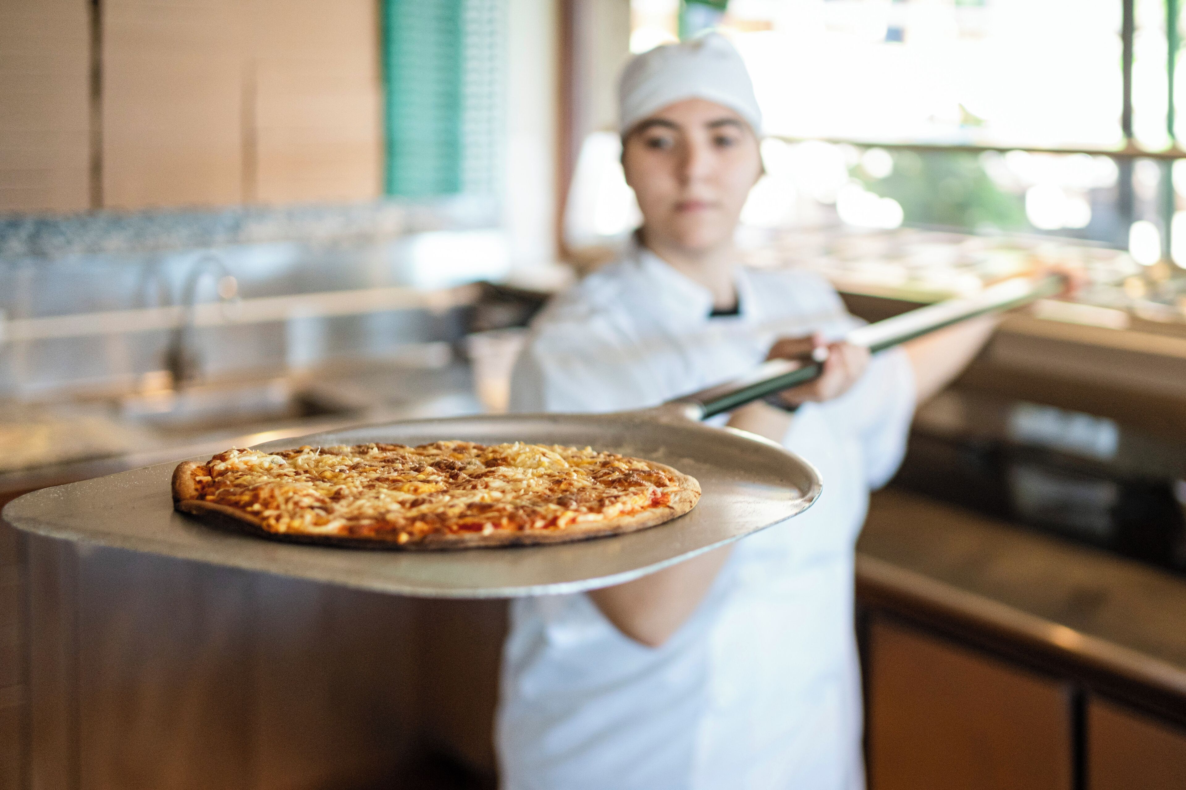 A chef holding a pizza on a metal tray in a kitchen.