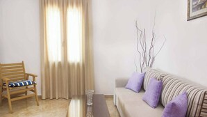 A living room with a wooden chair, a sofa with striped cushions, a glass vase on a table, and a window with curtains. - Villa Elena Glysteri (Skopelos)