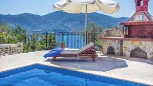 A pool area with a lounge chair and umbrella, a stone barbecue, and a view of mountains and the sea. - Villa Elena Glysteri (Skopelos)
