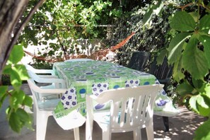 Una mesa con mantel floral, rodeada de sillas de plástico blanco, ubicada en un área exterior sombreada con follaje y un muro de piedra al fondo.