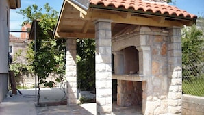 Estructura de horno exterior de piedra con tejado de azulejos, rodeada por una base de piedra y una zona pavimentada.