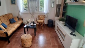 Una sala de estar con un sofá, un taburete redondo, una mesita con un florero, una silla de mimbre y un televisor sobre un mueble.
