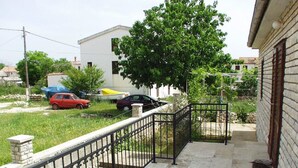 Un balcón con piso de piedra, baranda de metal y vista a coches estacionados y un árbol.