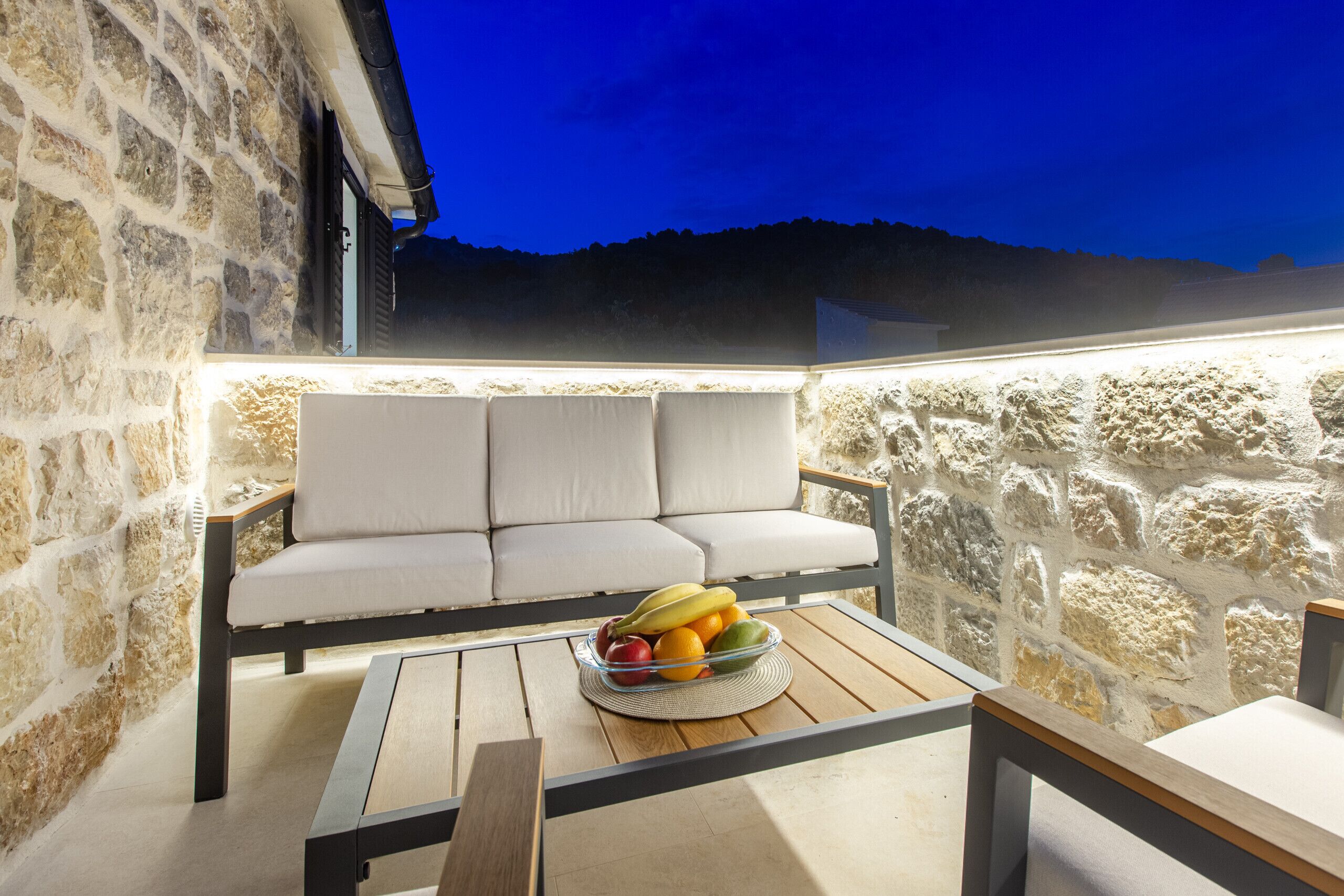 A stone wall, a cushioned bench, and a table with a fruit bowl.