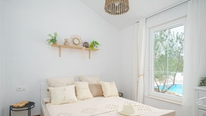 Een slaapkamer met een bed, nachtkastje, raam met uitzicht op een zwembad en een plank met decoratieve objecten.