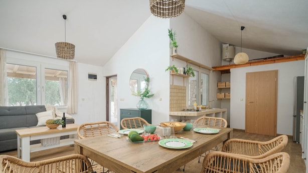 Een eethoek met een houten tafel gedekt voor vier personen, omringd door rieten stoelen. Er is een bank en een bijzettafel met een ananas en een fles erop.