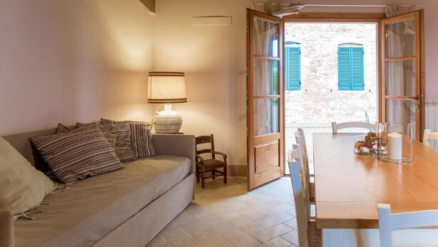 A cozy living room with a sofa, a table, and a chair. There is a lamp on the table and a window with blue shutters. - Apartment with 2 bedrooms and 2 bathrooms on the ground floor (Toscana)
