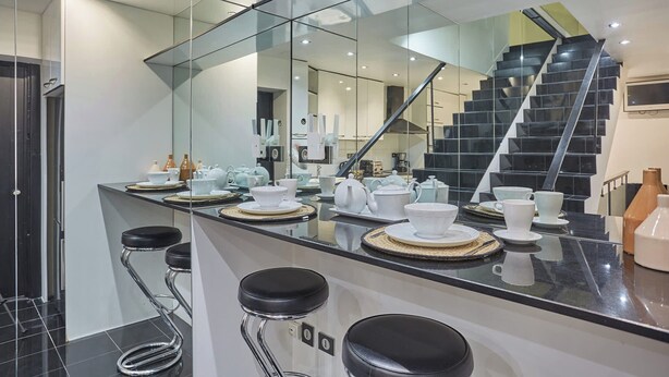 A modern kitchen with a glass backsplash, a stainless steel island with bar stools, and a staircase leading to an upper level. - Appartement Centre de Paris Le Marais (Paris)