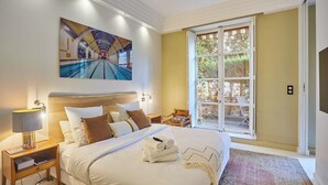 A hotel room with a large bed, a painting of a swimming pool, a window with a view of greenery, and a small table with a lamp. - Appartement Centre de Paris Le Marais (Paris)