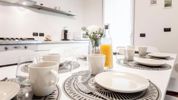 A modern kitchen with white cabinets, a stainless steel range hood, and a dining area with a white table set for four. - Bright & Modern 2-Bedroom Apartment in a Quiet Location (Milano)