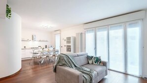 A modern living room with a sofa, a dining table, and chairs. There is a kitchen area in the background with cabinets and a countertop. - Bright & Modern 2-Bedroom Apartment in a Quiet Location (Milano)