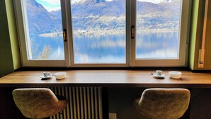 Une pièce dotée d’une grande fenêtre donnant sur un lac et des montagnes. On y trouve deux chaises et une table avec des tasses et des soucoupes.