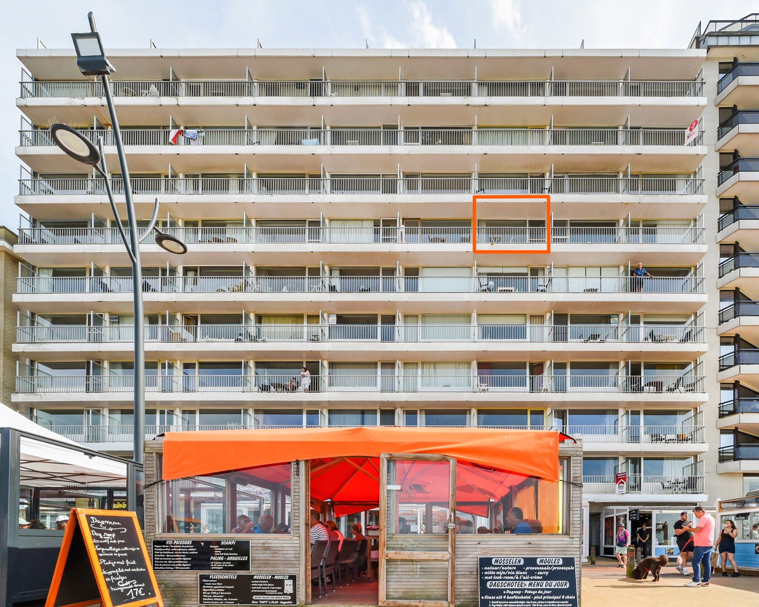 A multi-story building with balconies, a restaurant with an orange awning, and a signboard with menu items.