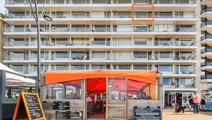 Un edificio de varios pisos con balcones, un restaurante con toldo naranja y un letrero con los platos del menú.