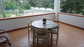 Un balcón con mesa y sillas, una planta en maceta y vistas a una cancha de tenis y árboles.