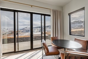 A modern room with a large sliding glass door leading to a balcony, a round table with chairs, and a view of snow-covered mountains.
