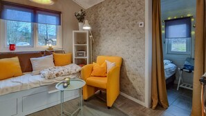 Een gezellige woonkamer met een bank, een klein tafeltje en een gele fauteuil. Er is een boekenplank en een raam met gordijnen.