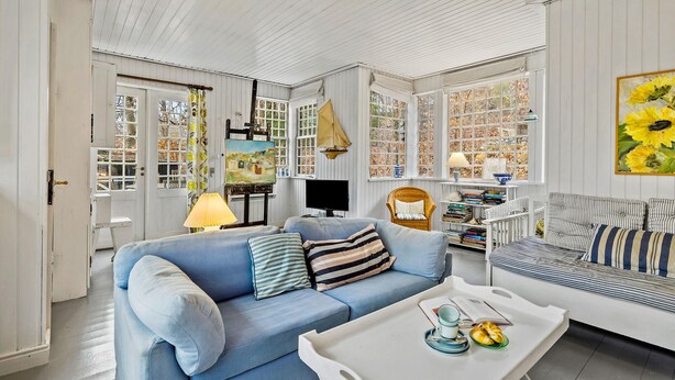 Ein Wohnzimmer mit einem blauen Sofa, einem weißen Couchtisch und einem gestreiften Sofa. An der Wand hängt ein Gemälde von Sonnenblumen, es gibt eine Segelboot-Dekoration und ein Bücherregal mit Büchern.