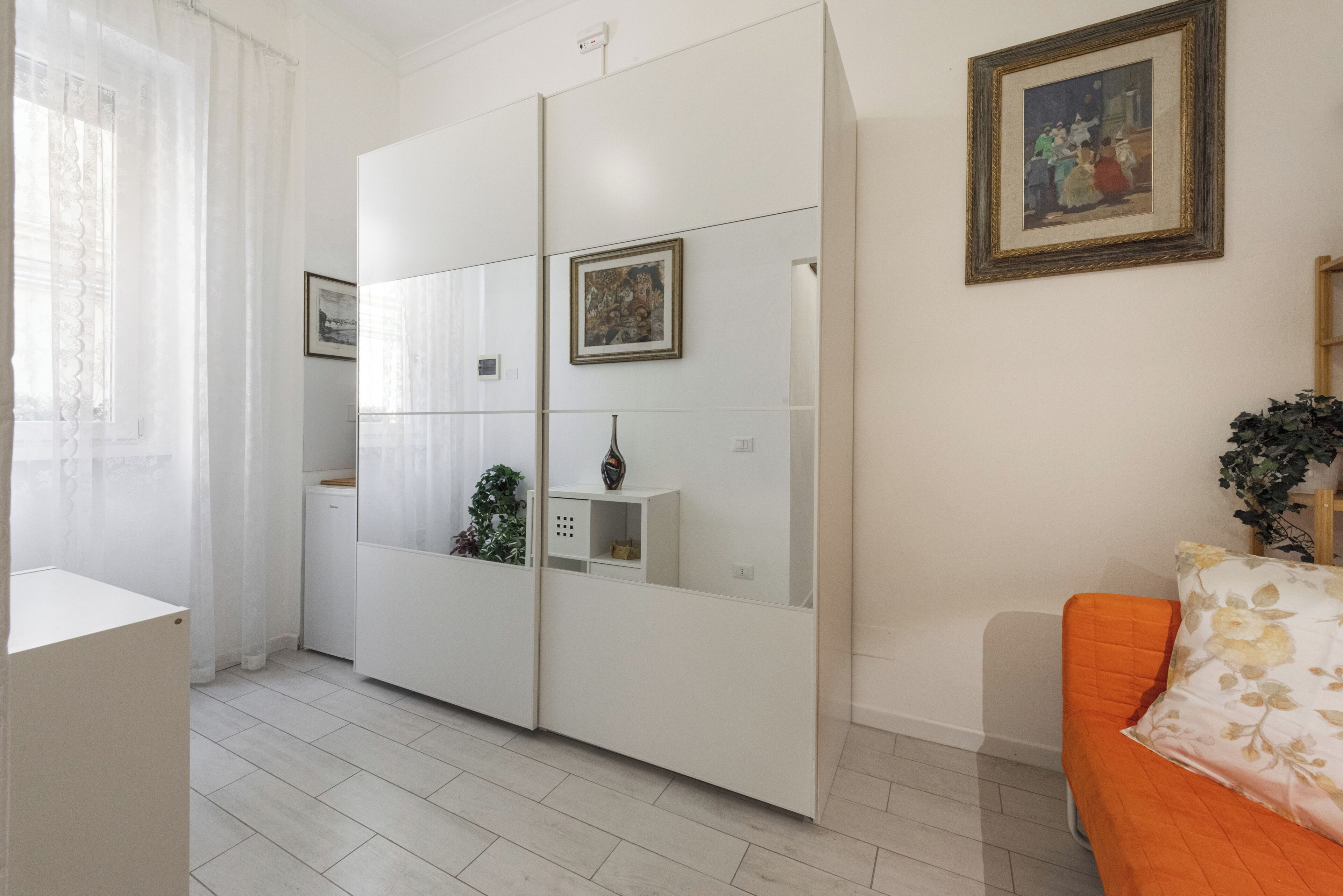 A room with a white wardrobe, a small plant, a framed picture on the wall, and a sofa with a floral cushion.
