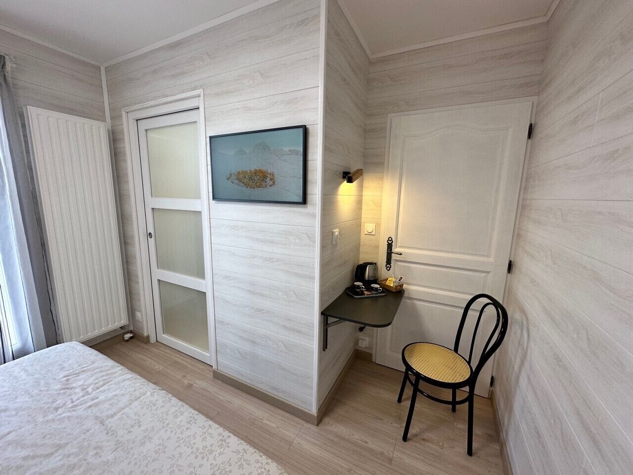 Une chambre à coucher avec un lit, un bureau avec une chaise, un tableau accroché au mur et une porte avec une lumière au-dessus.