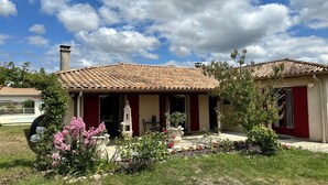 Une maison avec un toit en tuiles, une cheminée et un jardin agrémenté de fleurs roses et d’un kiosque blanc.