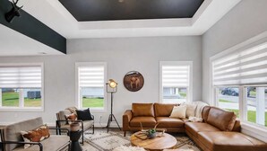 A modern living room with a sectional sofa, a coffee table, and a floor lamp. There are large windows with blinds, a ceiling with a black accent, and a decorative wall piece. - Modern rustic home near Bardstown top distilleries (Bardstown)