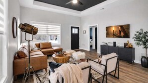 A modern living room with a ceiling fan, a large window with blinds, a sofa, a coffee table, and a television. - Modern rustic home near Bardstown top distilleries (Bardstown)