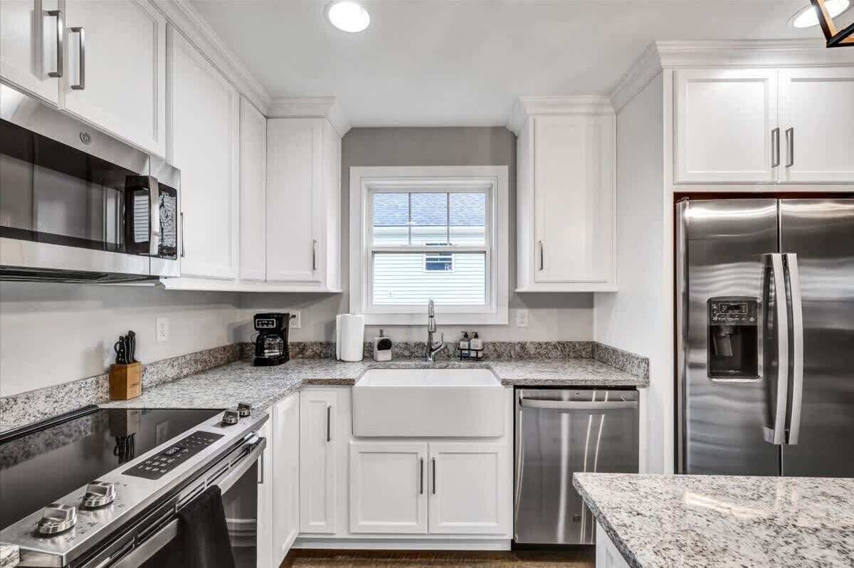 A modern kitchen with white cabinetry, stainless steel appliances, a granite countertop, and a window with blinds.