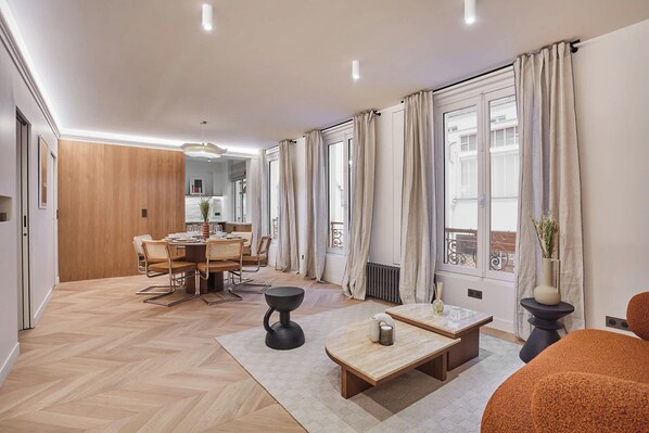 Een moderne woonkamer met een eethoek, grote ramen voorzien van gordijnen, en een houten vloer.