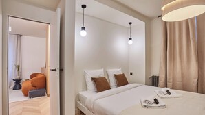 Une chambre d’hôtel moderne avec un grand lit, deux lampes de chevet, un petit fauteuil orange et une vue sur le couloir.