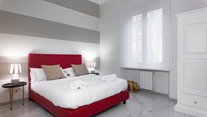 Een moderne hotelkamer met een rood bed, witte beddengoed, twee bedlampen, een gouden beeldje en een groot raam met luxaflex.