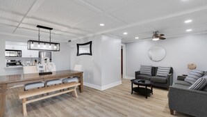 A modern living room with a wooden dining table, grey sofas, and a ceiling fan.