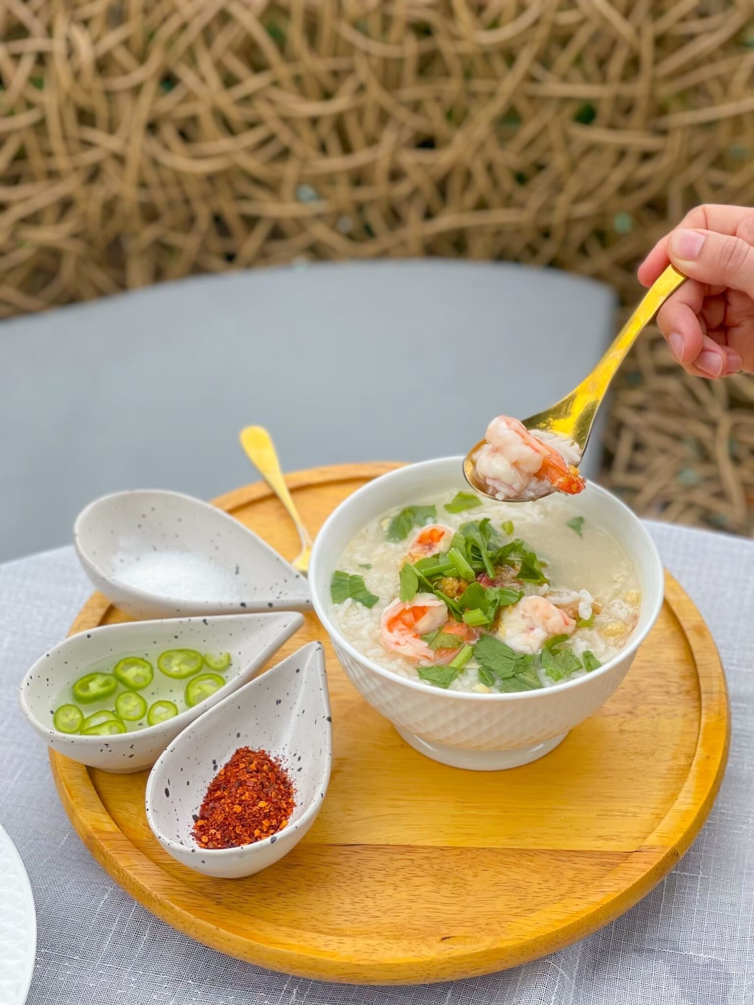 A bowl of soup with shrimp and herbs, accompanied by two small bowls containing sliced green chilies and red chili flakes on a wooden tray.