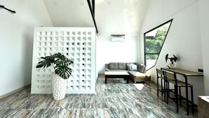 A modern, minimalist room with a geometric white partition, a large potted plant, a grey sofa, and a dining area with a wooden table and chairs. - The Estretto Resort (Wiang Chiang Rung)